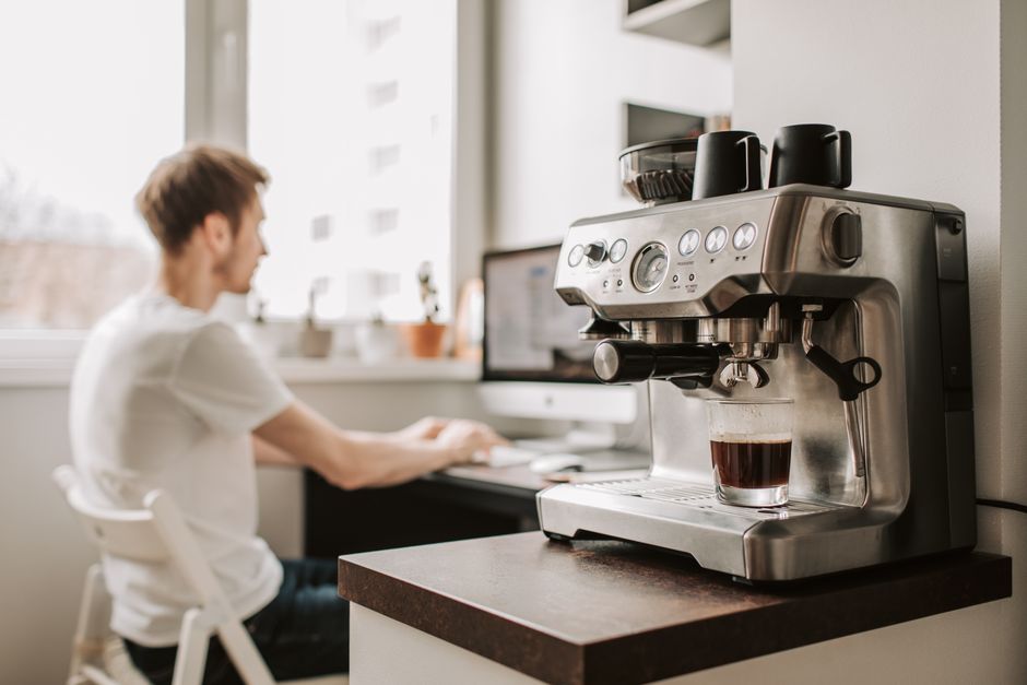 Side view of male remote worker in t shirt typing on desktop computer while sitting on plastic chair near window and coffeemaker in flat
