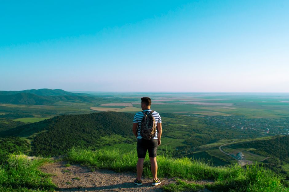 A man stands on a hilltop, overlooking a vast green landscape under a clear blue sky.