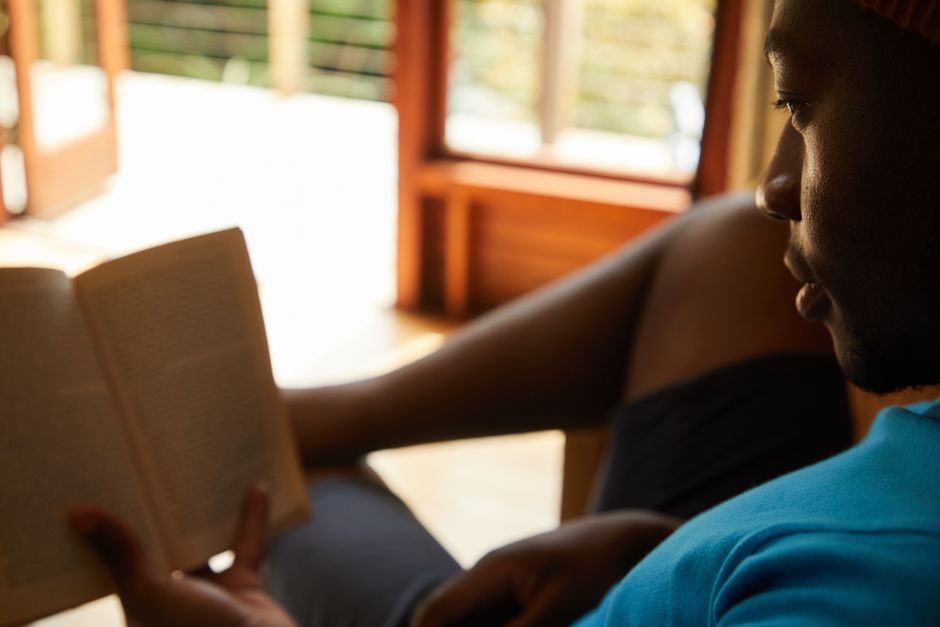 Crop African American male in casual clothes sitting on sofa and reading book while resting at home